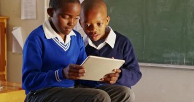 Ön planda, ilkokul 4k sınıfındaki derslere ara verilirken sandalyelerin arkasında oturan ve tablet bilgisayara bakan iki Afrikalı çocuğun görüntüleri var.