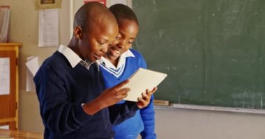 Bir ilkokul 4k sınıfındaki derslere ara verilirken sandalyelerin arkasında oturup tablet bilgisayara bakan iki Afrikalı çocuğun yan görüntüsü.