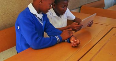 Bir ilkokul 4k sınıfındaki derslere ara verilirken masalarında oturup tablet bilgisayara bakan iki Afrikalı çocuğun yan görüntüsü.