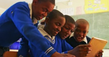 Bir ilkokul 4k sınıfındaki derslere ara verilirken dört Afrikalı çocuğun oturup tablet bilgisayara baktığı yan görüntü.