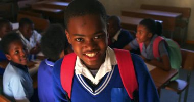 Okul üniforması ve okul çantası giymiş, kameraya bakıp gülümseyen Afrikalı bir çocuğun portresi. Arka planda sınıf arkadaşları oturuyor.
