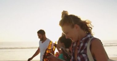 Gün batımında sahilde yürüyen, bira şişeleri içen ve serin bir kutu ve plaj çantaları taşıyan çok ırklı genç yetişkin arkadaşlardan oluşan çok ırklı bir grubun yan görünümüne yakın. Arka -dan aydınlatmalı. Arkadaşlarla yaz eğlencesi, onlar plaj 4k bir yolculuğa vardır