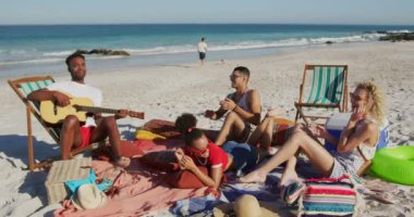 Mutlu genç yetişkin erkek ve kadın arkadaşlar dan oluşan çok ırklı bir grup, birlikte bir plajda oturan genç bir Afro-Amerikan adam şarkı ve gitar çalarken dinlerken. Genç arkadaşlar birlikte 4k plajda yaz eğlencesi