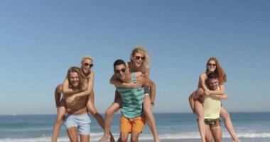 Bir plajda eğlenceli piggyback yarış altı mutlu genç yetişkin arkadaş çok ırklı bir grup yakın. Genç arkadaşlar birlikte 4k plajda yaz eğlencesi