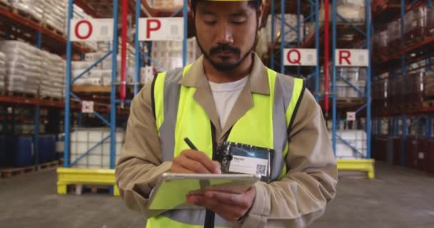 Gros plan d'un jeune ouvrier asiatique d'entrepôt portant un casque jaune sur un presse-papiers debout dans une baie de chargement d'entrepôt. Storarge étagères sont visibles en arrière-plan . 