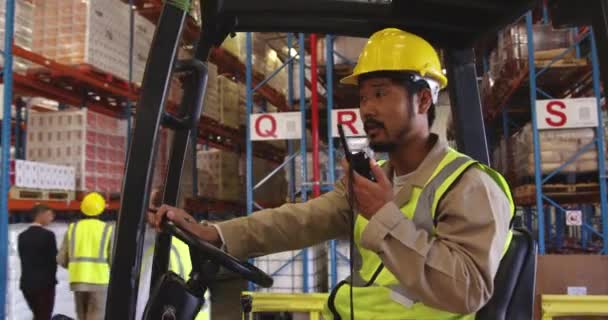 一名年轻的亚洲男性仓库工人坐在叉车上 戴着一顶黄色硬礼帽 在仓库装卸库中使用双向无线电 仓库的工作人员在仓库的架子上走来走去 — 图库视频影像