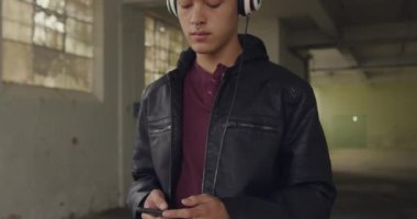 Boş bir depoda bir kalça genç karışık Yarış adam Ön görünümü, kulaklık ve gözleri kapalı müzik dinleyen bir akıllı telefon tutarak