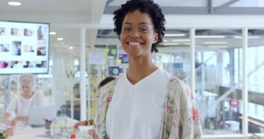 Modern açık planlı ofiste kameraya gülümseyen genç, melez, yaratıcı bir kadının portresi.