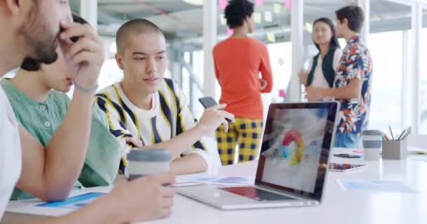 Vue de côté gros plan d'un groupe de jeunes professionnels de la création parlant autour d'un ordinateur portable dans un bureau moderne, avec des collègues parlant en arrière-plan 