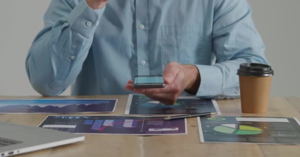 スマートフォンを使ってテーブルに座っている若い白人男性の正面図の中間部分で ラップトップコンピュータ テイクアウトコーヒーをテーブルの上に置きます — ストック動画