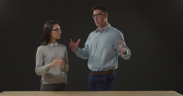 黒い背景のワークベンチの後ろに立つ議論で保護安全眼鏡を身に着けている若い白人男性と女性の正面図クローズアップ — ストック動画