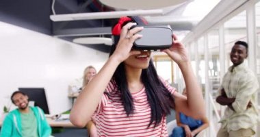 Genç, melez, yaratıcı, VR google 'lar takan, etrafa bakınan ve çeşitli meslektaşları tarafından izlenen modern, açık planlı bir ofisteki genç bir kadının ön görüntüsü.
