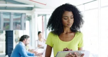 Genç, melez, yaratıcı bir kadının tablet bilgisayar kullanan ve arka planda iş arkadaşlarının konuştuğu modern bir açık plan ofisinde gülümseyen kameraya bakan portresi.