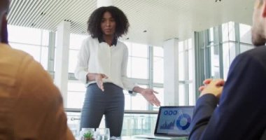 Genç bir Afrikalı Amerikalı iş kadınının modern bir ofiste sırtı kameraya dönük iki erkek meslektaşına dizüstü bilgisayar kullanarak sunumunu yapması ön planda.