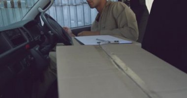 Bir minibüsün sürüş koltuğunda oturan genç bir karışık ırk erkek depo işçisi nin ön görünümü yakın, bir pano ve kalem ve yazma, bir depo dışında ulaşan