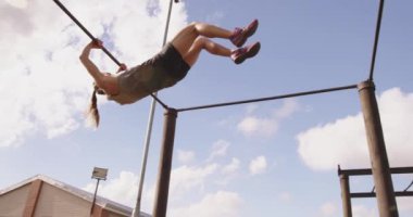 Bir acemi birliği eğitim seansı sırasında, açık hava spor salonunda, parmaklıklar arasında sallanan ve zıplayan beyaz bir kadının yan görüntüsü.