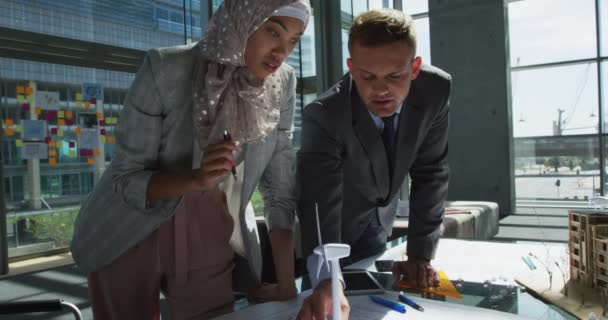 一位身穿头巾的年轻混血女商人和一位年轻的白人商人站在桌旁 看着一台模特儿风力涡轮机 在一个现代办公室的会议上交谈 — 图库视频影像