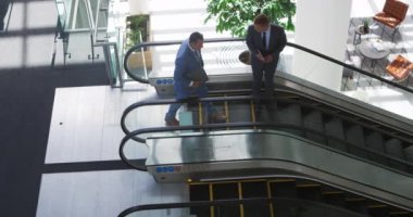 Kafkasyalı genç bir iş adamı ve orta yaşlı beyaz bir işadamının modern bir ofis binasında yürüyen merdivenden düşerken konuşurken görüntüsü yüksek. Biri dizüstü bilgisayar, diğeri tablet bilgisayar tutuyordu.