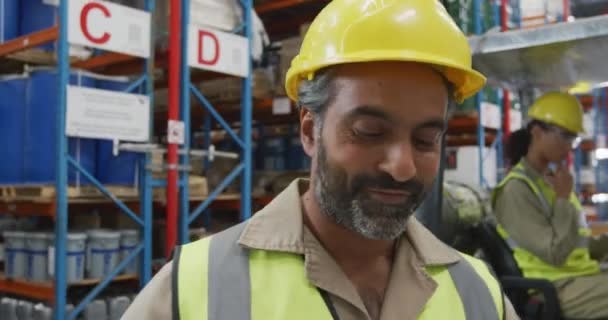 倉庫荷室でカメラに笑顔黄色のハード帽子を身に着けている中年の混合レース男性倉庫労働者の肖像画は 背景にフォークリフトトラックの同僚と — ストック動画