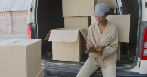 一个年轻的混血女仓库工人坐在一辆装满箱子的敞车里 在仓库装卸区外的剪贴板上写字 — 图库视频影像