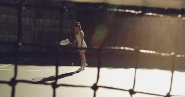 Kortta tenis oynayan, servise hazırlanan topu zıplatan, bir ağdan görülen beyaz bir kadının ön görünümü