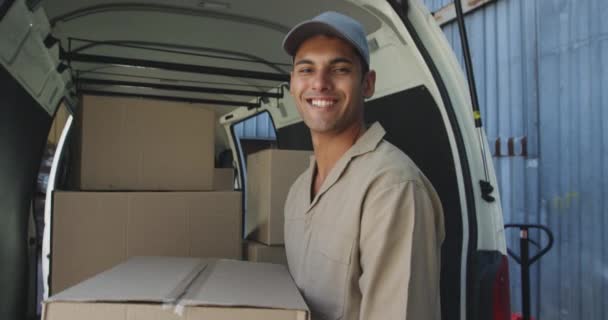 一个年轻的混血男仓库工人拿着一个盒子 微笑着站在仓库外一辆装满箱子的敞开货车的后面 对着镜头微笑 — 图库视频影像
