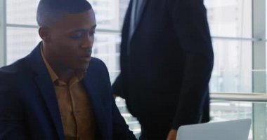 Genç bir Afro-Amerikalı iş adamının yakın çekimde dizüstü bilgisayarla oturmuş arkasında duran Kafkasyalı genç bir işadamıyla modern bir ofiste bilgisayar ekranına bakıp konuşurken ve gülümserken.