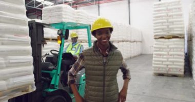 Portre yakın bir depo yükleme koyunda gülümseyerek kameraya yürüyen genç bir karışık ırk kadın depo yöneticisi, arka planda bir forklift kamyon üzerinde bir meslektaşı ile