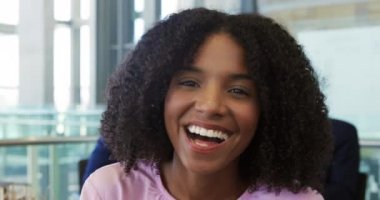 Kameraya gülümseyen ve modern bir ofiste kahkahalarla gülen genç bir Afrikalı Amerikalı iş kadınının portresi. Arka planda çalışan bir erkek meslektaşıyla birlikte.