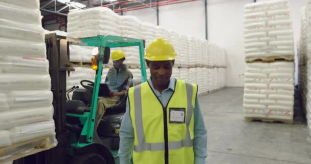 Portrait rapproché d'un homme d'âge moyen gérant d'entrepôt mixte marchant vers la caméra souriant dans une baie de chargement de l'entrepôt, avec un collègue sur un chariot élévateur à fourche en arrière-plan 