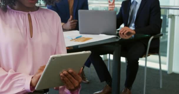 一个年轻的非洲裔美国女商人在办公室里用平板电脑做特写 眼睛望着相机笑着 两个男同事坐在书桌前 背景是一台笔记本电脑 — 图库视频影像