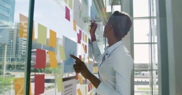 一位年轻的非洲裔美国女商人 一位年轻的混血女商人站在玻璃墙边 用彩色粘贴的便条在一间现代化的办公室里 侧观近景 — 图库视频影像