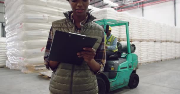 Portrait fermer un jeune gestionnaire d'entrepôt mixte écrit sur un presse-papiers souriant à la caméra dans une baie de chargement de l'entrepôt, avec un collègue sur un chariot élévateur à fourche en arrière-plan 