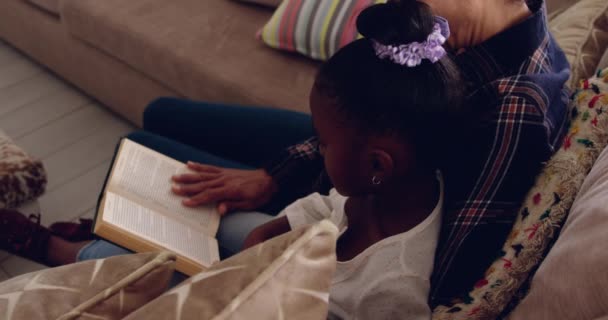 高角度的看法 一个成熟的混血女人坐在她的小孙女读一本书与她在家里 — 图库视频影像