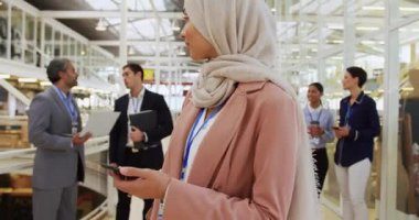 Asya 'lı genç bir iş kadınının portresi tesettür takıyor, akıllı bir telefon tutuyor, kameraya bakıyor ve gülümsüyor, bir grup farklı iş adamı antredeki bir iş konferansında arka planda konuşuyor.