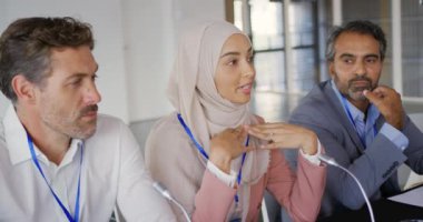 Üç iş adamı, beyaz bir adam, tesettür takan genç bir Asyalı kadın ve bir iş konferansında önlerinde mikrofonlarla oturan orta yaşlı bir ırk adamı.. 