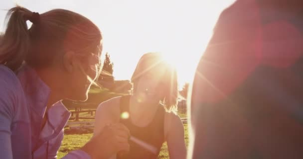 Par-dessus l'épaule d'une jeune femme caucasienne et d'un jeune homme caucasien qui écoutent une jeune entraîneuse caucasienne qui leur donne de la rétroaction assise sur l'herbe dans un gymnase extérieur lors d'une séance d'entraînement de bootcamp, rétroéclairée par la lumière du soleil 