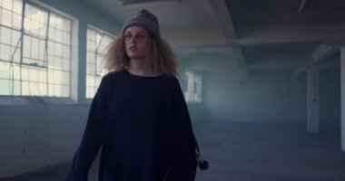 Boş bir depoda bir kalça genç Kafkas kadın ön görünümü, bir kaykay taşıyan ve kameraya yürüme