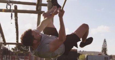 Arka planda diğer katılımcılarla birlikte acemi kampı antrenmanı sırasında, açık hava spor salonunda yatay ipe tırmanan, baş aşağı asılı genç bir beyaz adamın yan görüntüsü.