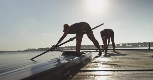 两个年轻的成年白种女赛艇运动员站在码头上准备桨和赛车壳在河上训练前的侧视图 — 图库视频影像