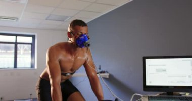 Eğitim sırasında metabolik gaz analizörü kullanan genç bir karışık ırk erkek bisikletçinin ön görünümü, yanında bilgisayar monitörü olan bir yüz maskesi takmak