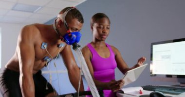 Genç bir karma ırk erkek bisikletçi eğitim sırasında bir metabolik gaz analizörü kullanarak Ön görünümü, genç bir Afro-Amerikan kadın kişisel eğitmen bir tablet tutan ve bir bilgisayar ekranında sonuçları kontrol ederken bir yüz maskesi giyiyor