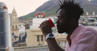 Çatının tepesindeki bir partide eğlenen, içen ve arka plandaki binaların manzarasına hayran olan mutlu Afrikalı genç bir Amerikalı 'nın yan görüntüsü.