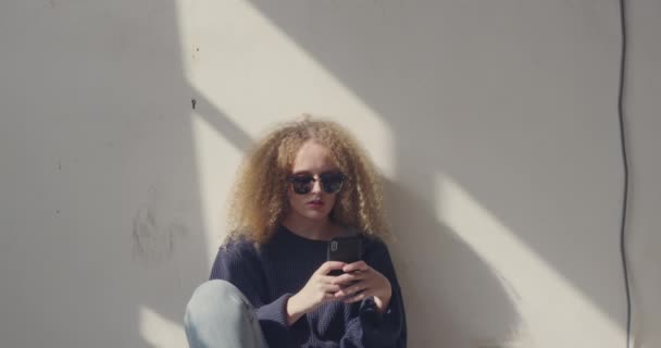 一个时髦的年轻白种女人在空仓库前视图 使用智能手机坐在墙上 — 图库视频影像