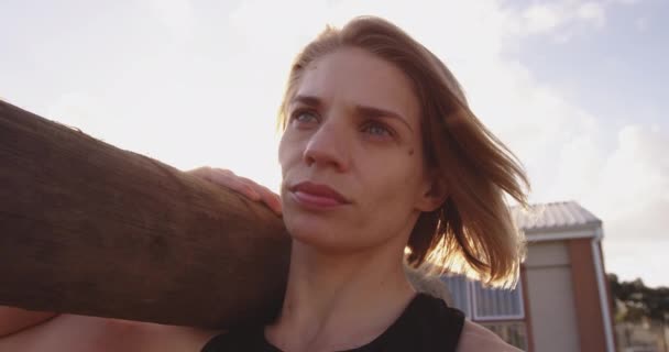 若い白人女性が肩に木の丸太を持っていて ブートキャンプのトレーニングセッション中に屋外のジムでカメラを探している肖像画 バックライト — ストック動画
