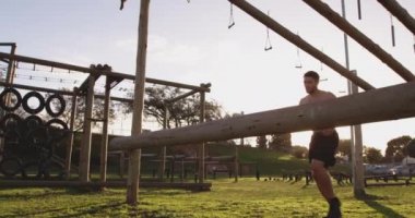 Bir acemi birliği eğitim seansı sırasında açık hava spor salonundaki bir engelin üzerinden atlayan üstsüz beyaz bir adamın yan görüntüsü.