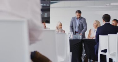 Orta yaşlı bir melez işadamının, bir iş konferansında oturan meslektaşları ve delegeleri tarafından çevrelenmiş bir seyirciye seslenişi. Seyircinin arkası ön planda görünür.