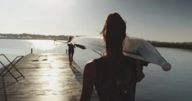 Nehirüzerinde eğitim almadan önce iskelede omuzlarında yarış mermisi taşıyan iki genç yetişkin Beyaz kadın kürekçinin arka dansı