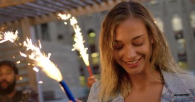 Genç, mutlu, beyaz bir kadının çatıdaki bir partide arka planda arkadaşlarıyla havai fişeklerle dans edişinin ön yüzü.