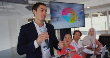 Beyaz gömlekli ve siyah ceketli beyaz tenli genç bir işadamının bir kürsüde dikilip üzerinde bilgi olan bir ekrana işaret ederken ve bir iş konferansında izleyicilere hitap etmek için mikrofon kullanırken yan görüntüsü.